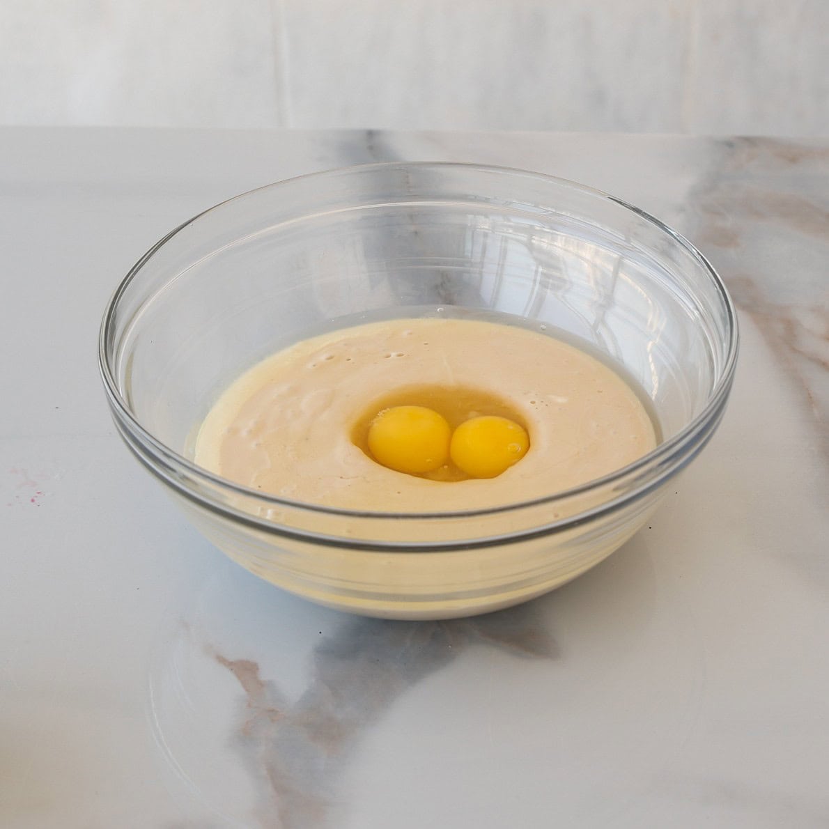 A glass bowl with muffin batter and two raw eggs sits on a white marble countertop.