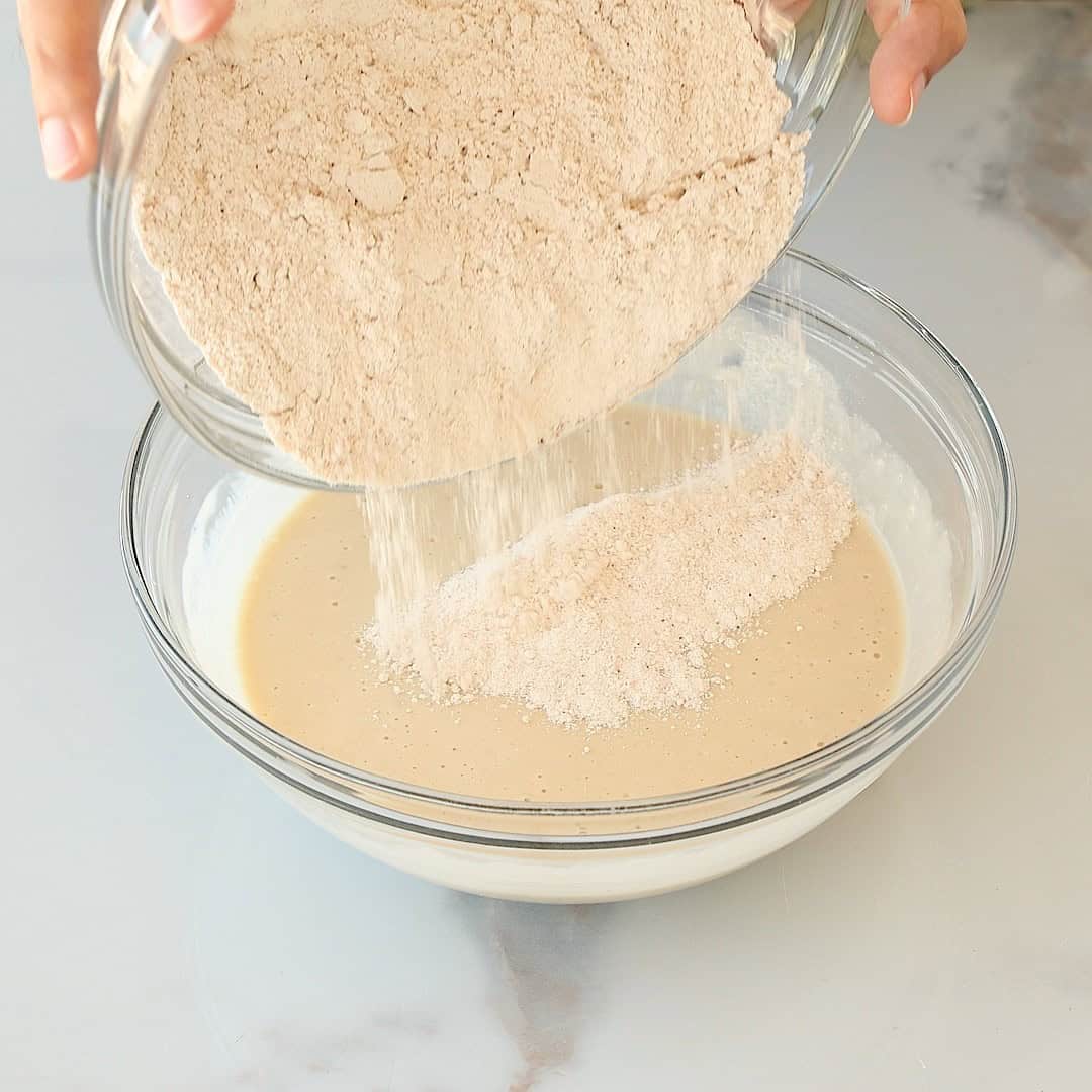 A person pours dry flour mixture from a glass bowl into a larger glass bowl containing a wet muffin batter on a light marble surface.