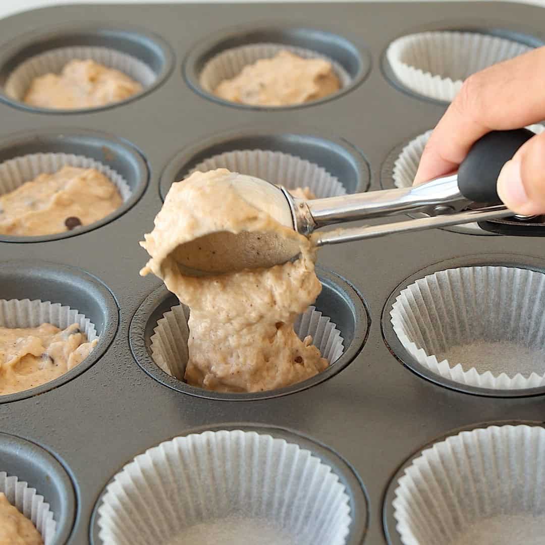 A hand uses an ice cream scoop to fill a lined muffin tin with thick banana cottage cheese muffin batter. Several cups are already filled, while others are empty and lined with paper.