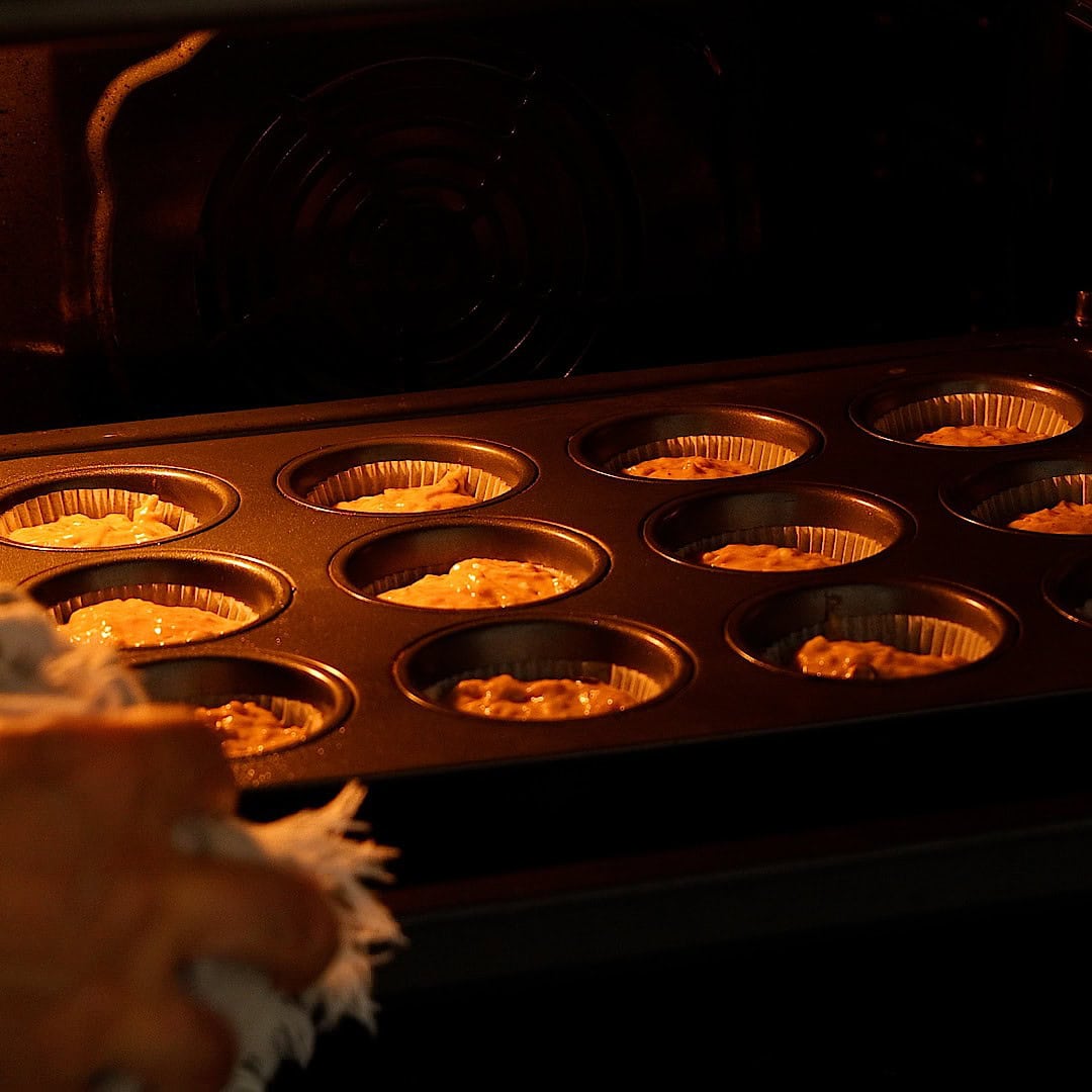 A person places a muffin tray filled with batter into an oven, preparing to bake cupcakes. The scene is warmly lit, highlighting the tray and the oven interior.