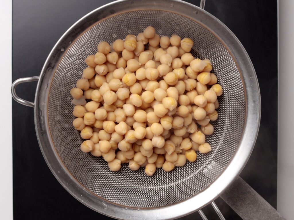 A metal strainer filled with cooked chickpeas is placed over a dark surface, with steam rising from below, suggesting the beans have just been drained.