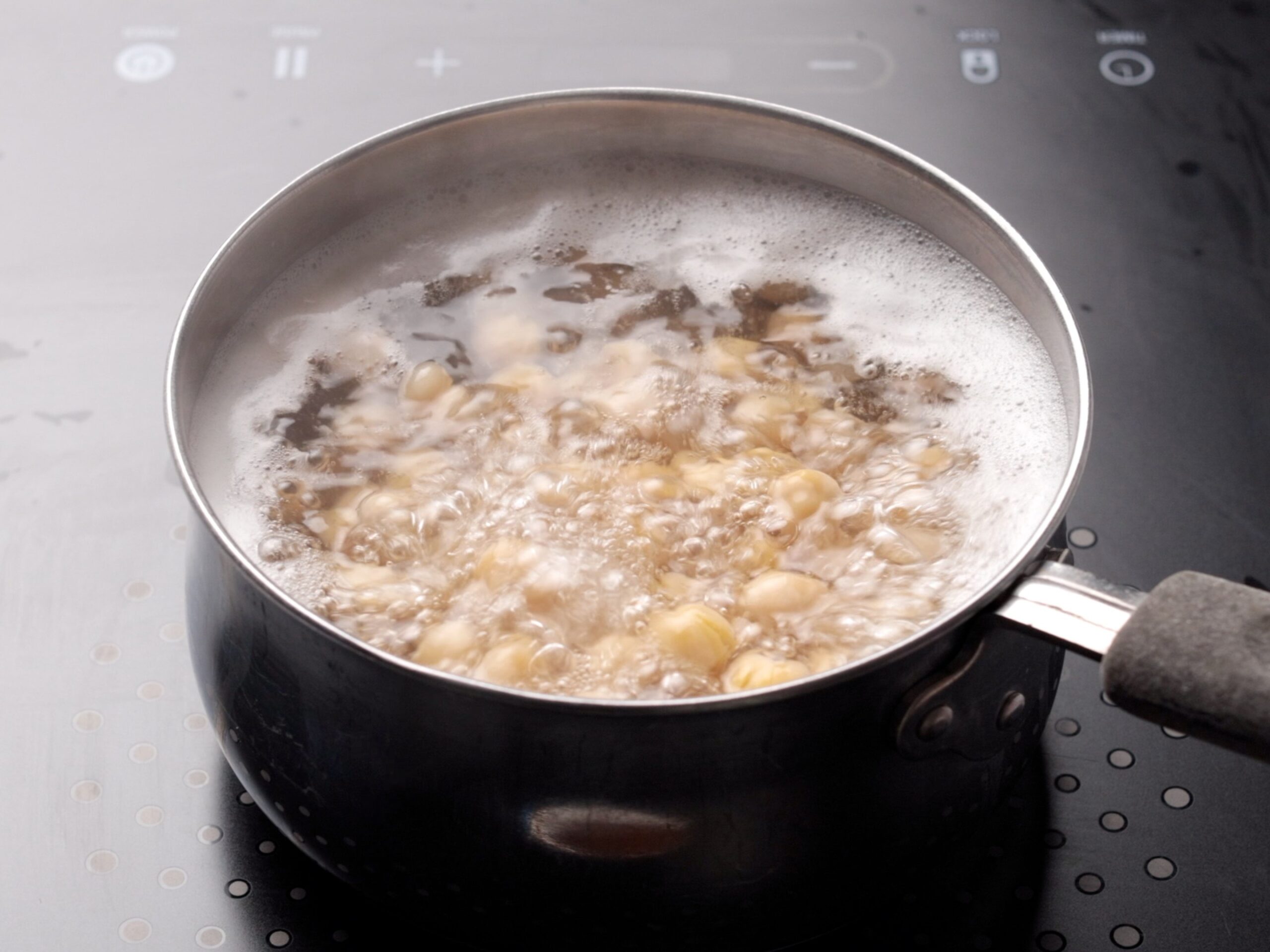 A saucepan of beans boiling in water sits on a stovetop with bubbles rising to the surface, showing active cooking in progress.