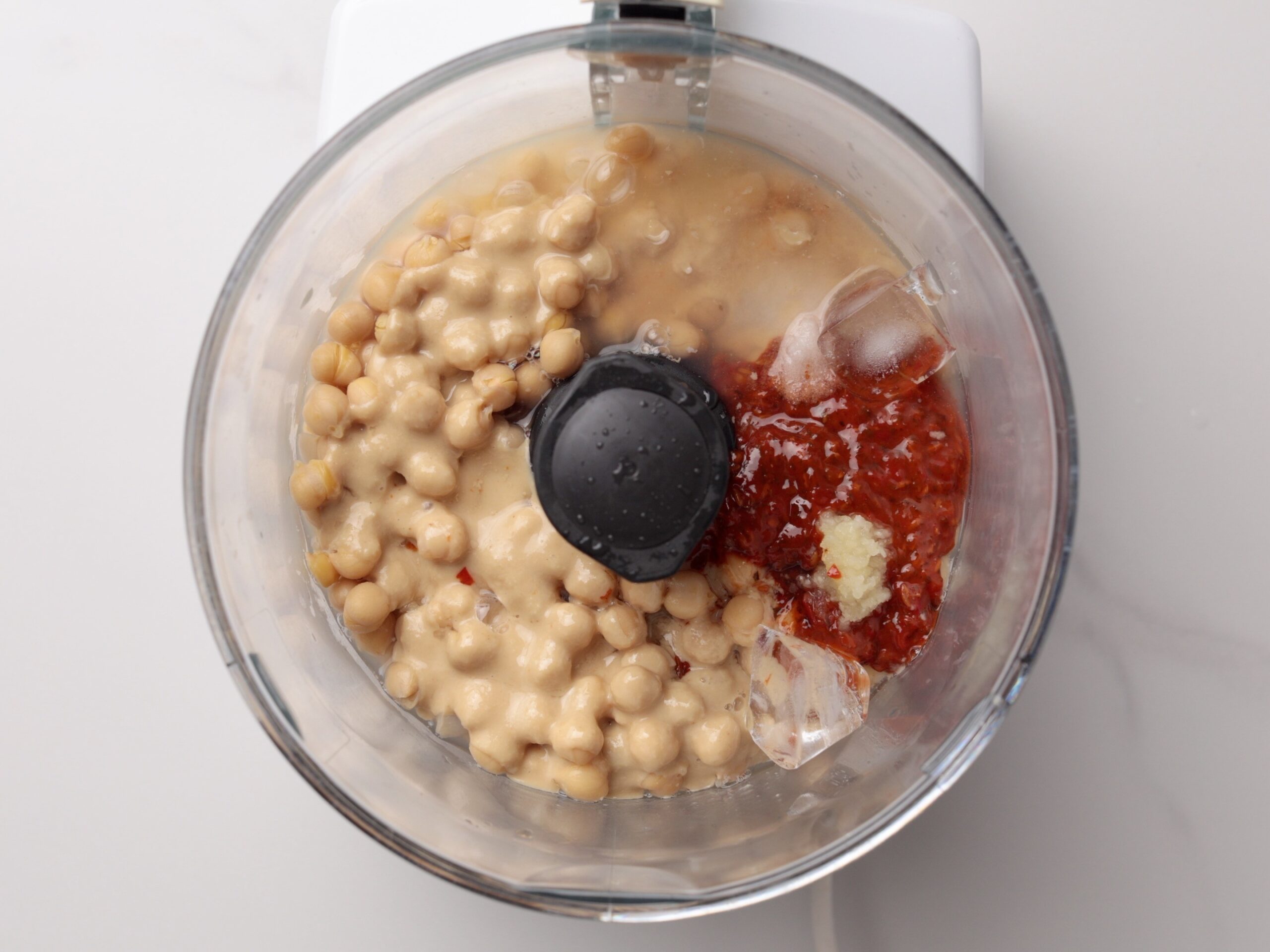 A food processor bowl filled with chickpeas, tahini, harissa paste, garlic, and ice cubes, ready to be blended, viewed from above on a white surface.