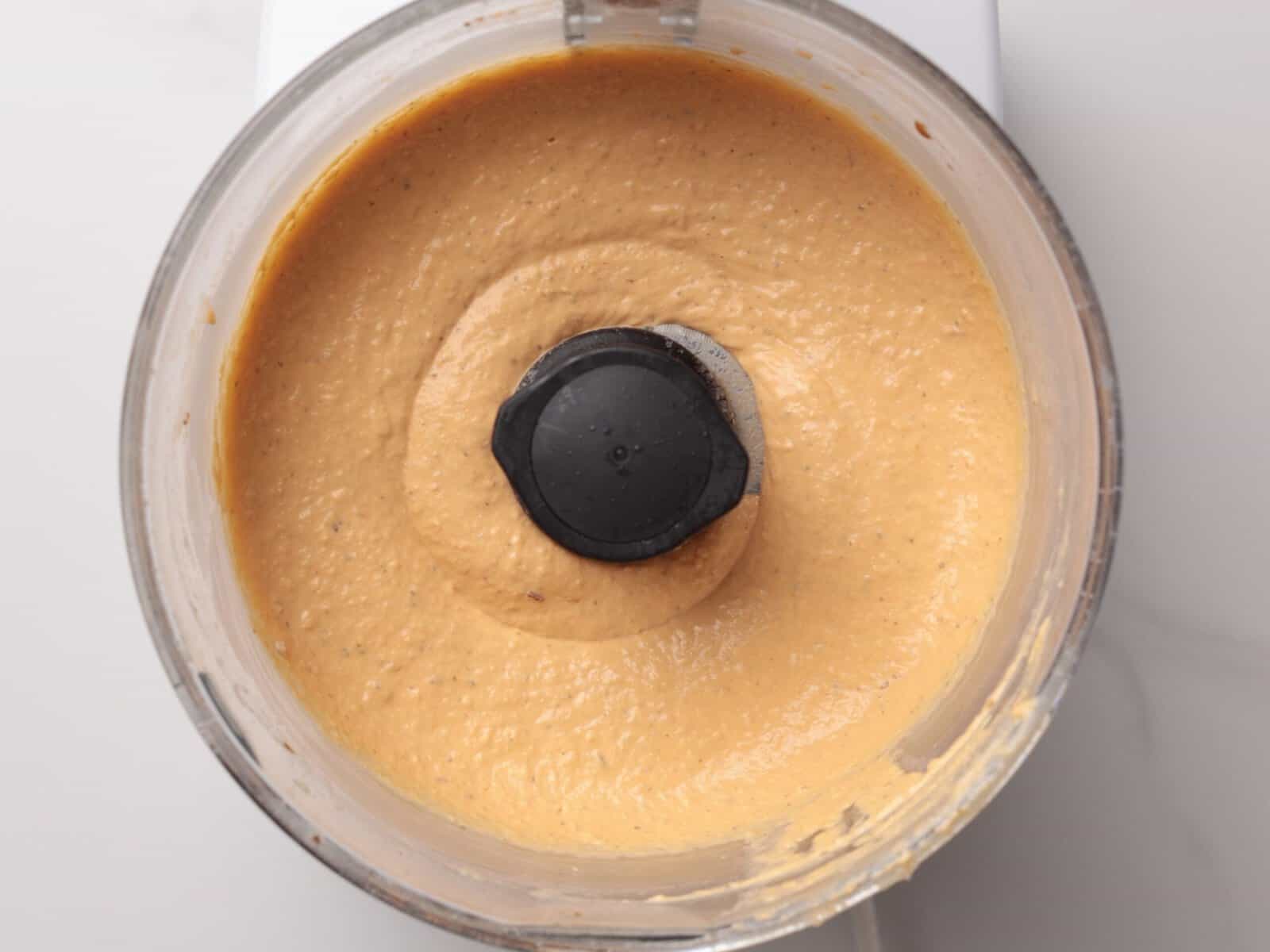 A food processor filled with creamy, light hummus with harissa, viewed from above, on a white countertop.