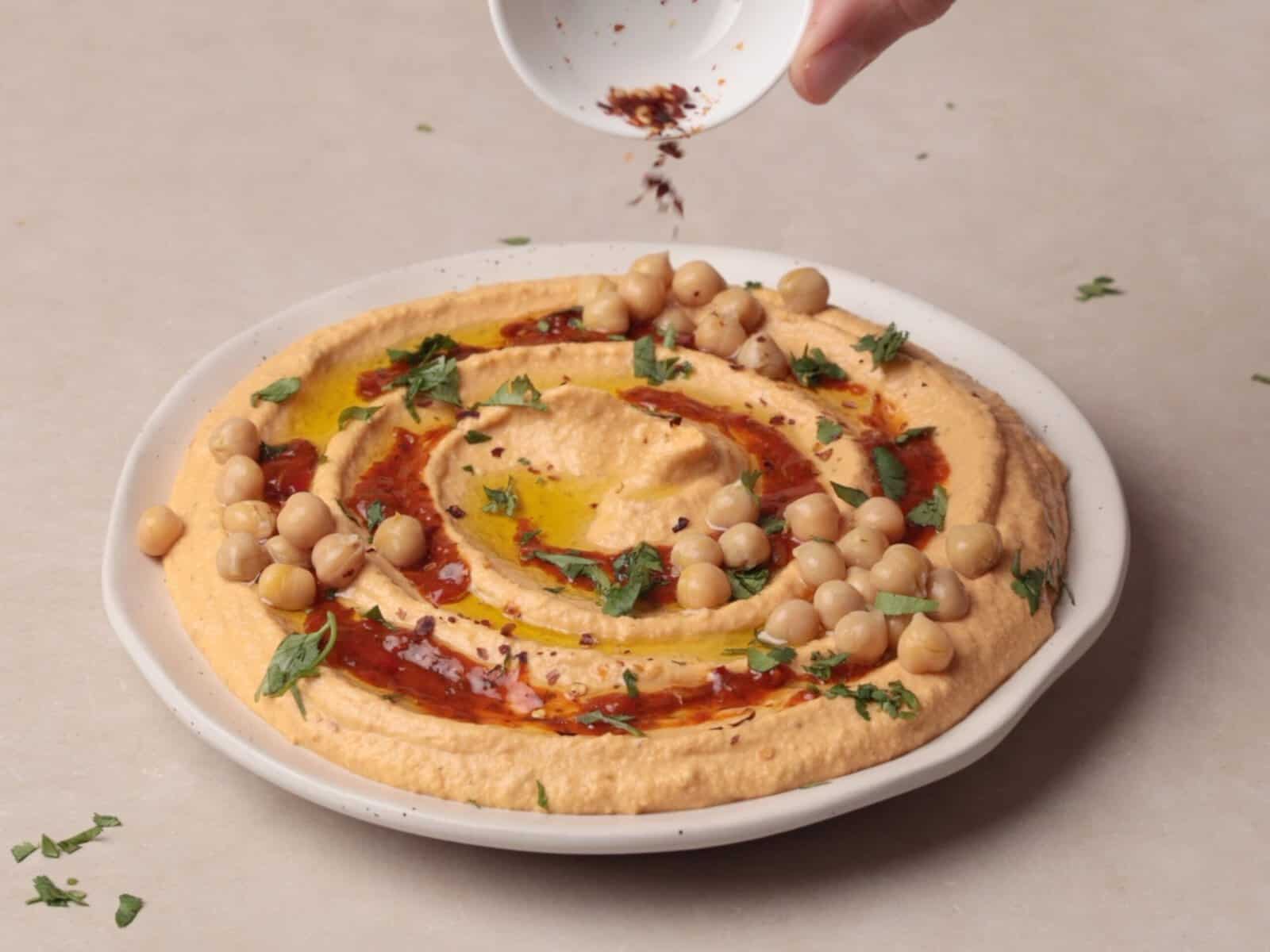 A hand sprinkles spices onto a plate of hummus harissa mix garnished with chickpeas, olive oil, herbs, and spices, presented on a light-colored surface.