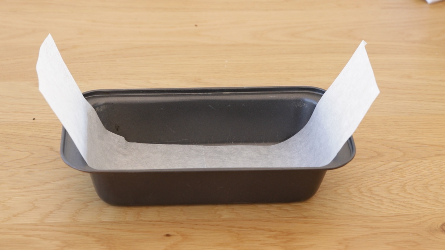 A rectangular black loaf pan sits on a wooden surface, lined with a sheet of parchment paper that extends above the two longer edges of the pan.