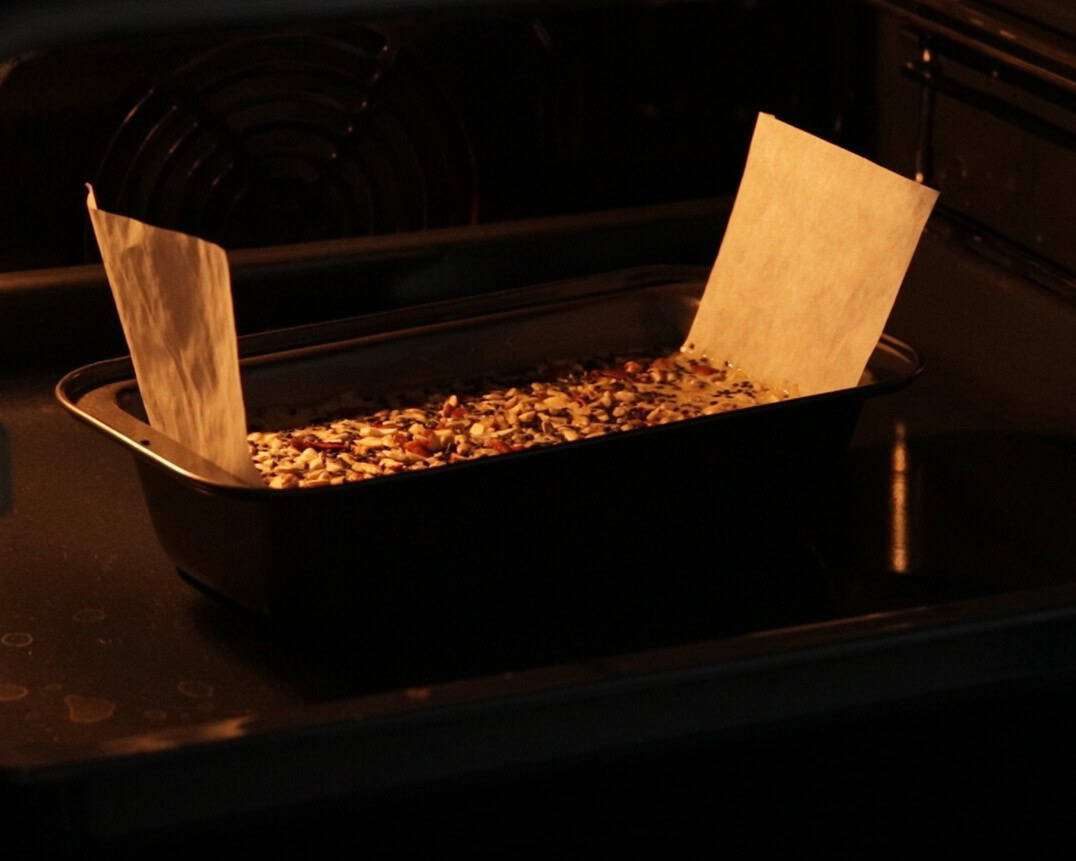 A loaf pan lined with parchment paper and filled with tahini bread topped with seeds and nuts is baking in an oven.