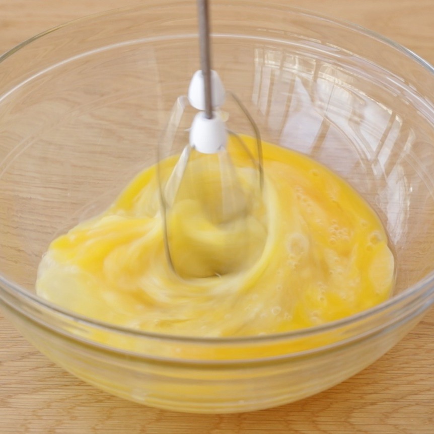 A handheld mixer beats yellow eggs in a clear glass bowl on a wooden surface, blending the mixture smoothly.