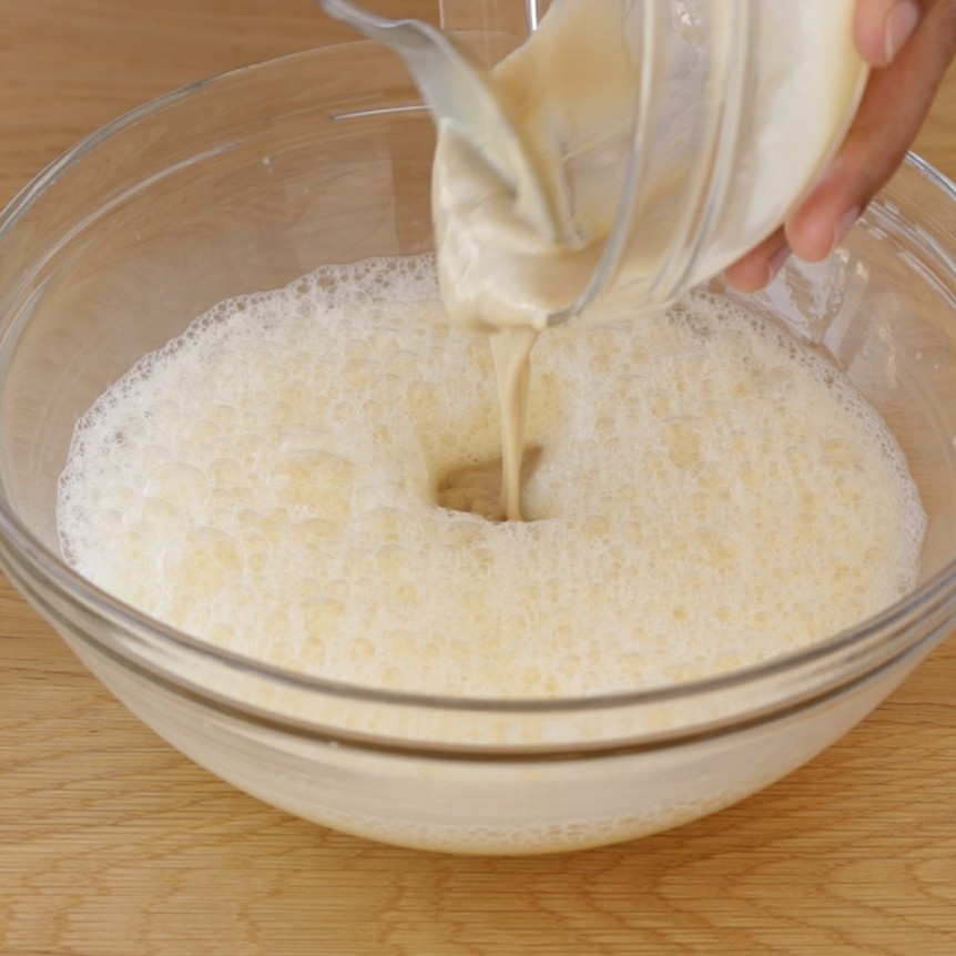 A hand pours a creamy tahini from a small bowl into a larger glass bowl filled with a frothy, bubbly mixture, all on a wooden surface.