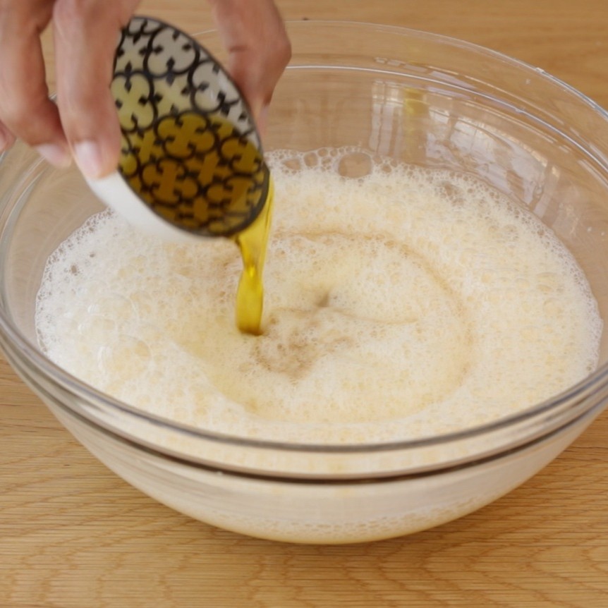 A hand pours olive oil from a small patterned bowl into a large glass bowl filled with a frothy mixture on a wooden surface.