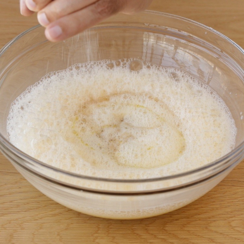 A hand sprinkles salt into a clear glass bowl filled with frothy, gluten-free bread batter on a wooden surface.