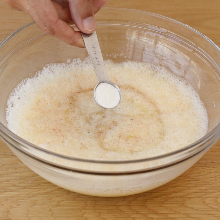 A hand adds a teaspoon of baking powder, likely baking soda, to a large glass bowl containing a frothy, low-carb tahini bread batter, on a wooden surface.