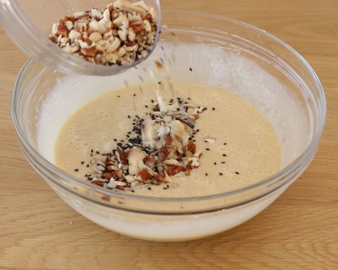 A hand pours chopped nuts and seeds into a bowl of a gluten free bread batter on a wooden table.