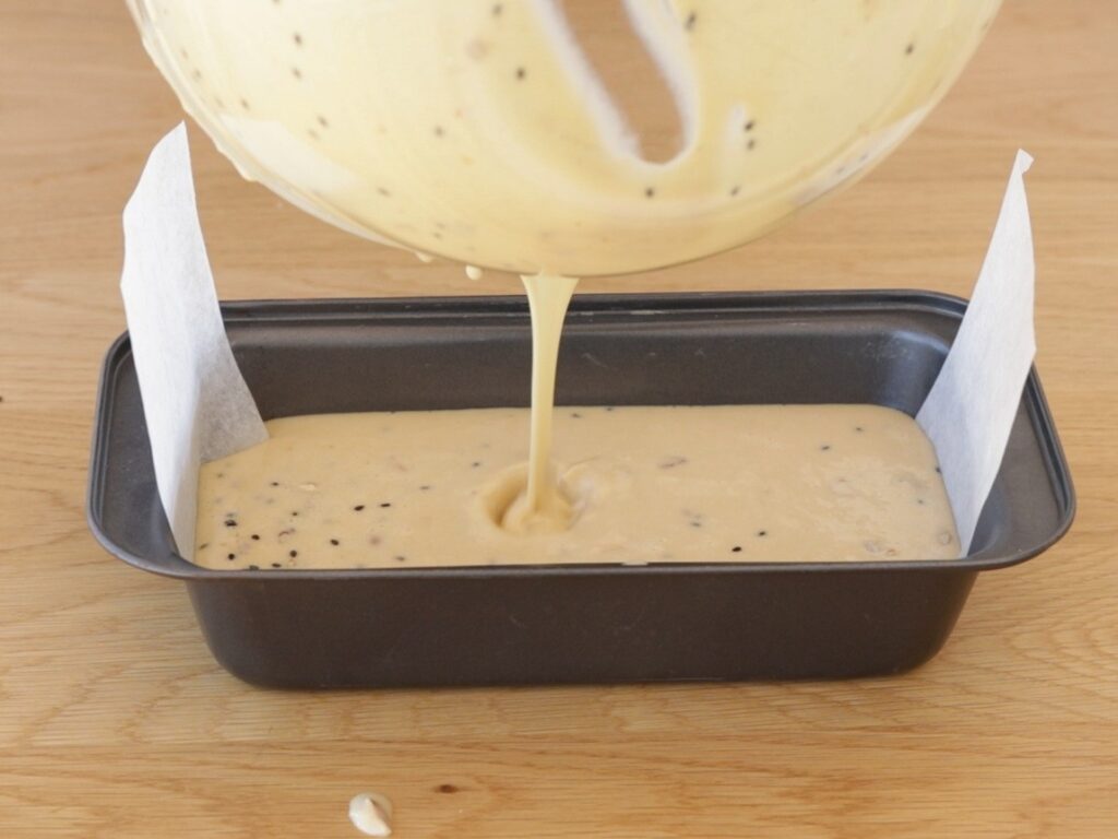 Thaini bread batter is being poured from a bowl into a lined loaf pan, ready for baking. Some batter has already filled the pan, and a drop has spilled onto the wooden surface.