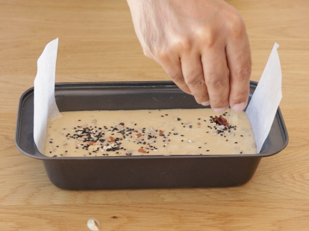 A hand sprinkles seeds or nuts onto a flourless low-carb bread batter in a parchment-lined loaf pan on a wooden surface.