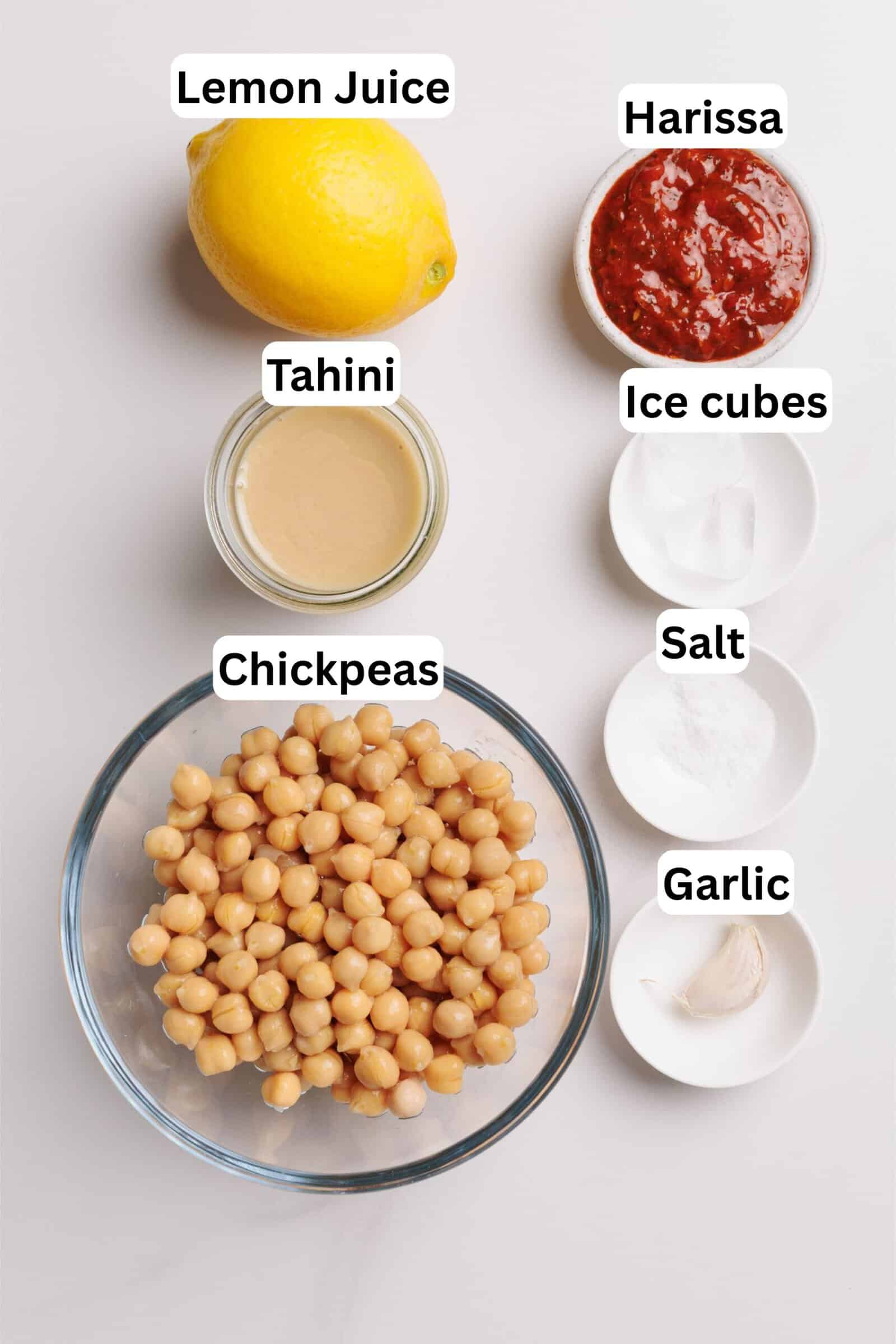 A bowl of chickpeas, a lemon, a jar of tahini, a bowl of harissa, salt, ice cubes, and a garlic clove are arranged on a white surface, each labeled with text.