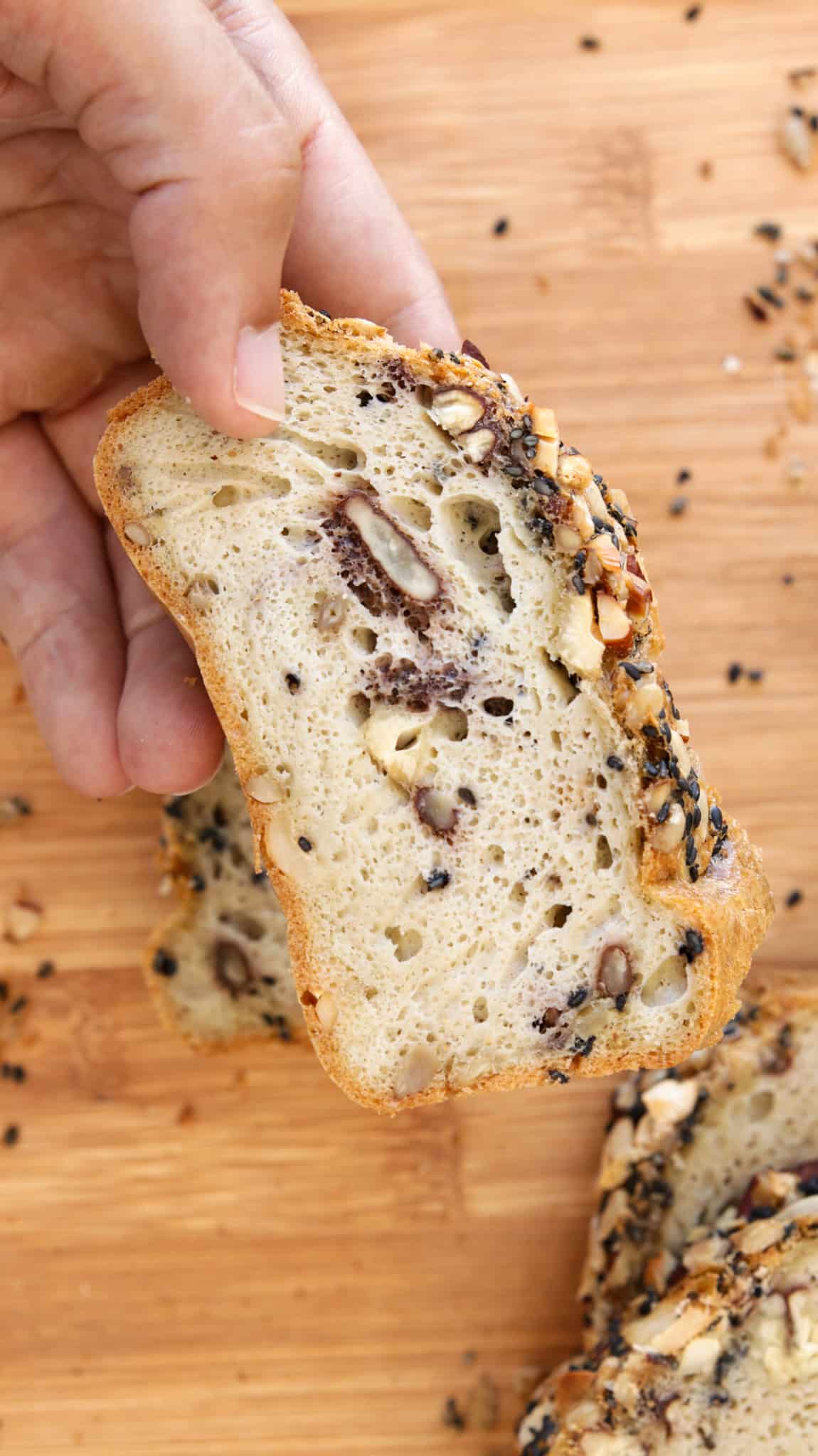 A hand holds a slice of gluten-free tahini bread with a light, airy crumb and visible nuts, above a wooden surface with scattered seeds and crumbs.