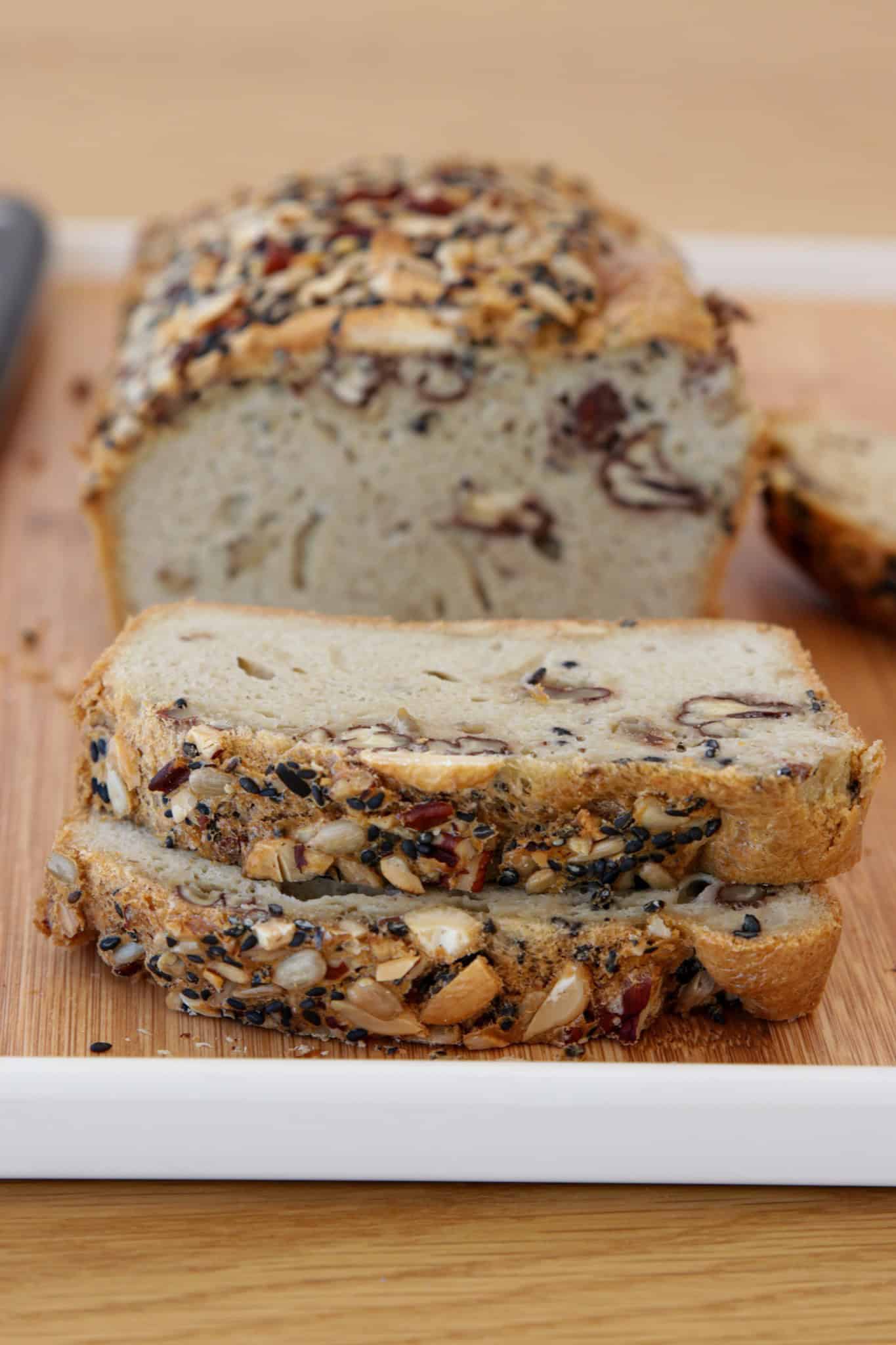 A loaf of multigrain gluten-free tahini bread with seeds and nuts on top is sliced on a wooden cutting board. Two slices are in the front, showing the texture and mix of ingredients inside.