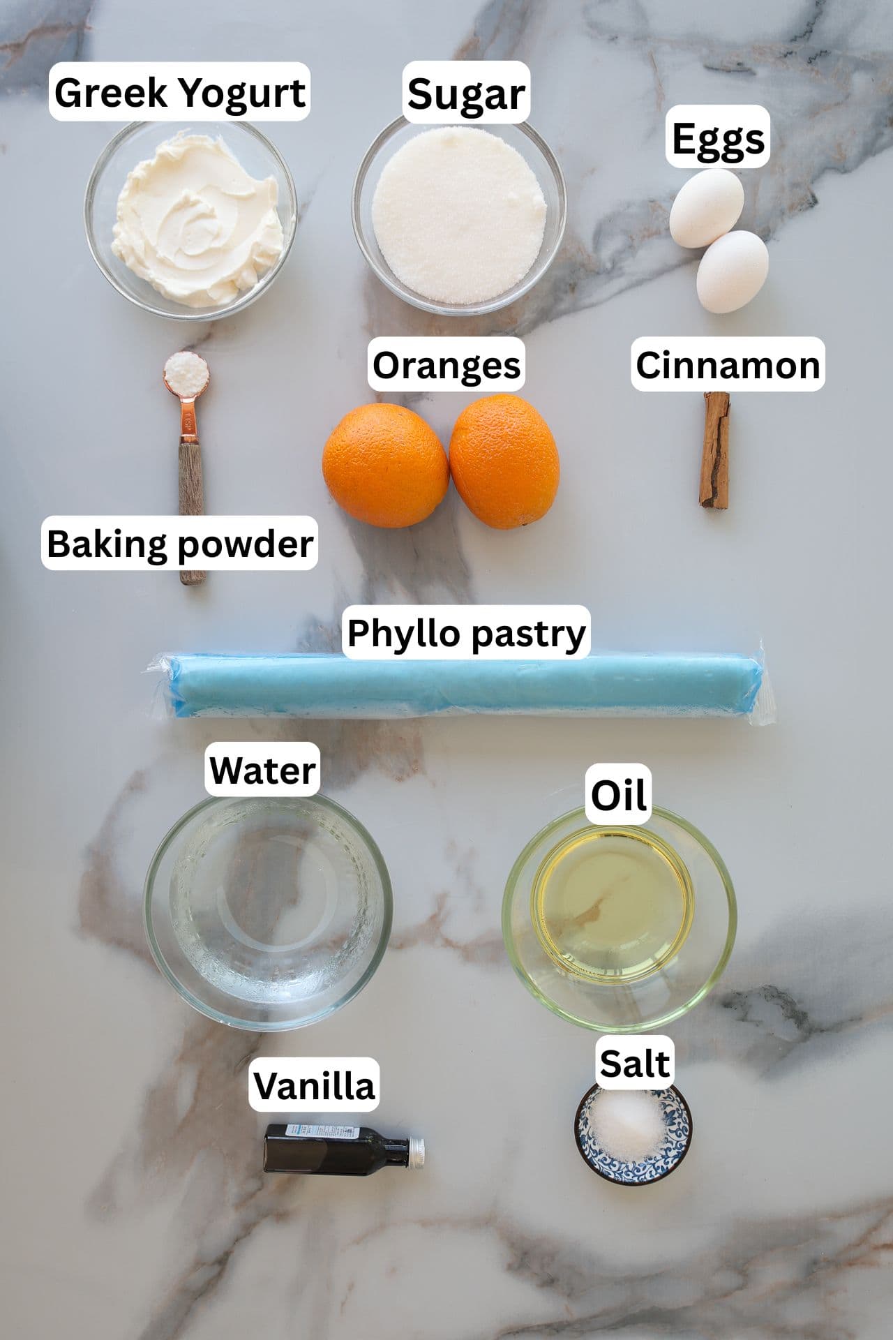 Ingredients for a recipe are arranged on a marble surface, including Greek yogurt, sugar, eggs, oranges, cinnamon, baking powder, phyllo pastry, water, oil, vanilla, and salt, each labeled clearly with text.