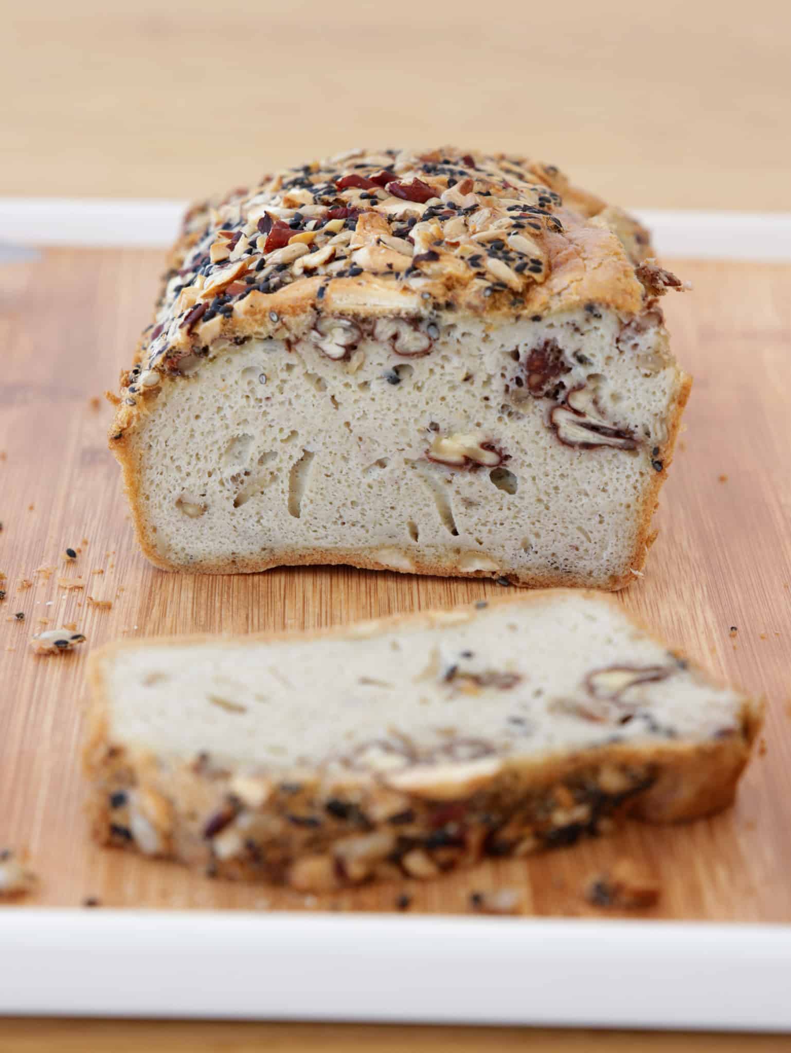 A tahini bread topped with seeds and nuts sits on a wooden cutting board, with one slice cut and placed in front of the loaf.