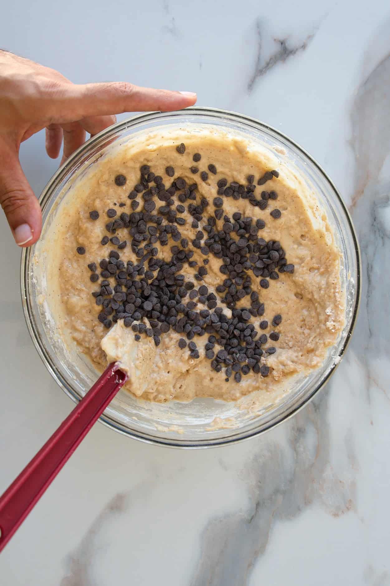 A hand holds a glass bowl of a healthy high-protein muffin batter with chocolate chips on top, being mixed with a red spatula on a white marble surface.