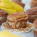 A banana muffin with a crinkled paper liner sits in front of whole bananas and other muffins, all on a light surface.