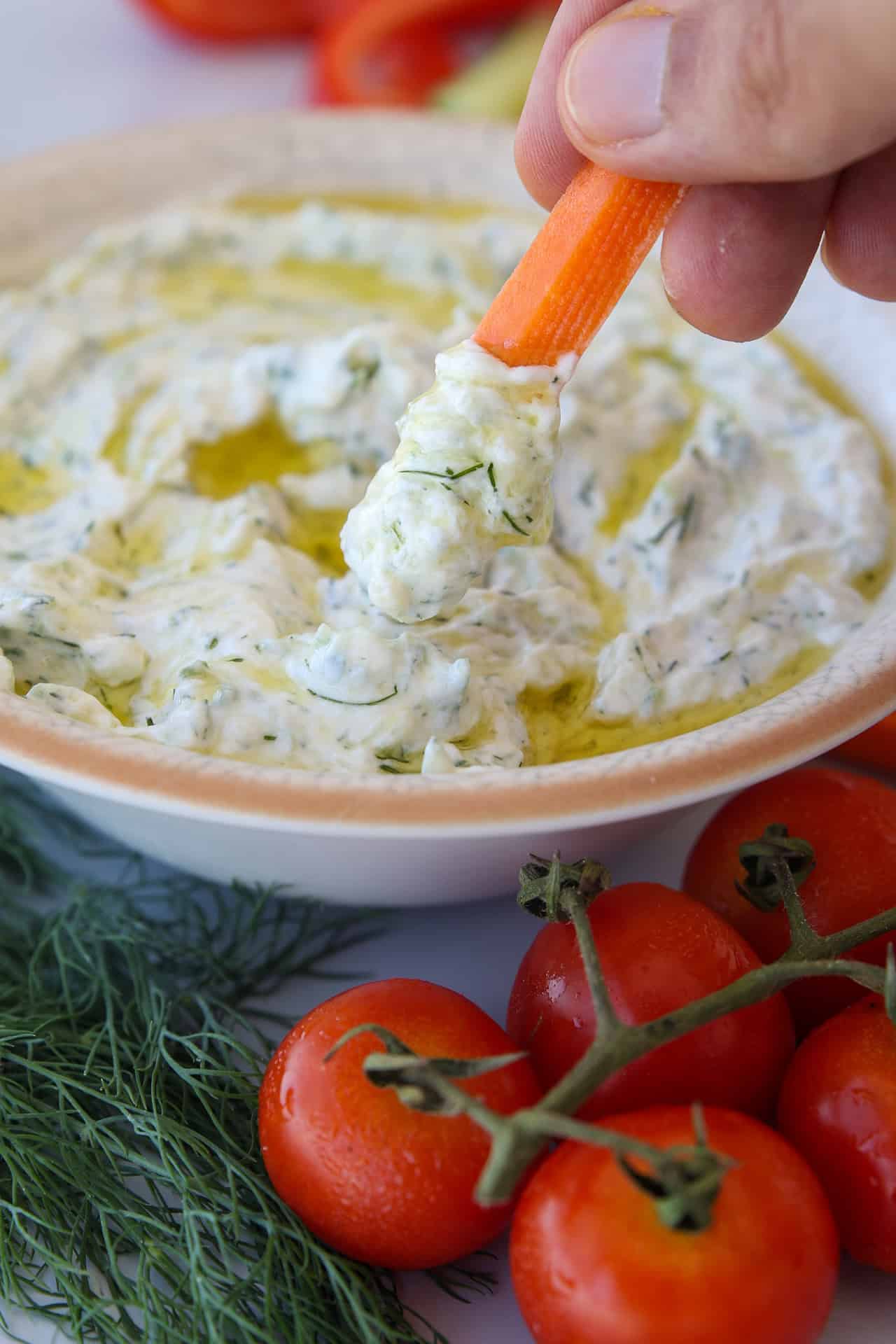 A hand dips a carrot stick into a creamy Greek tzatziki dip with herbs, with fresh dill and tomatoes on the table.
