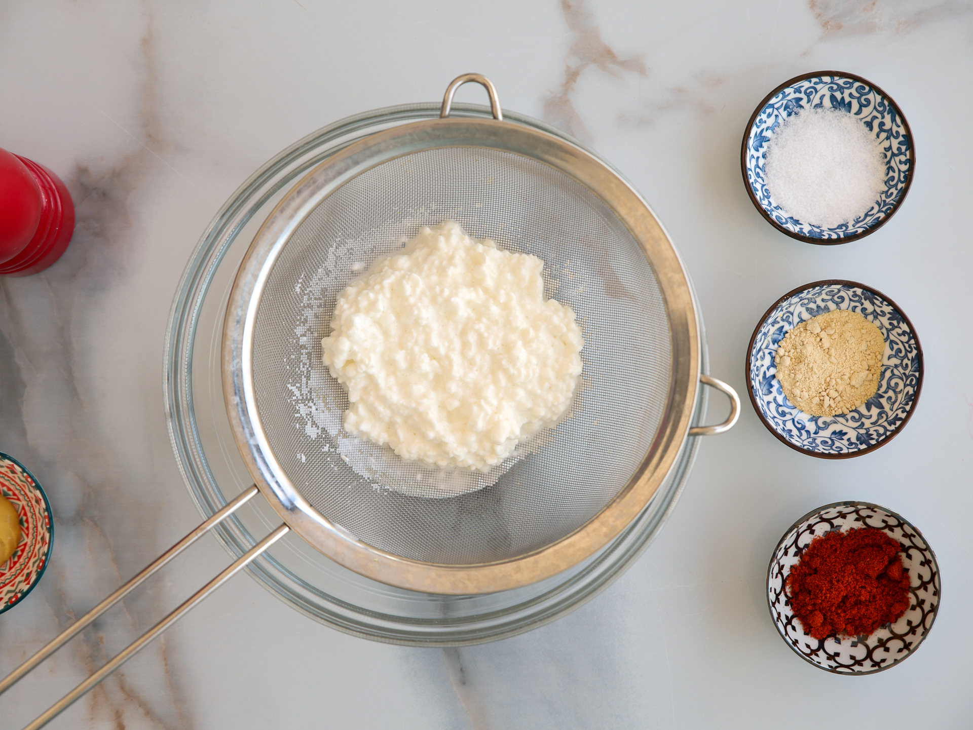 A metal strainer with cottage cheese sits over a glass bowl. Nearby are small bowls containing salt, garlic powder, and paprika, all on a marble surface.