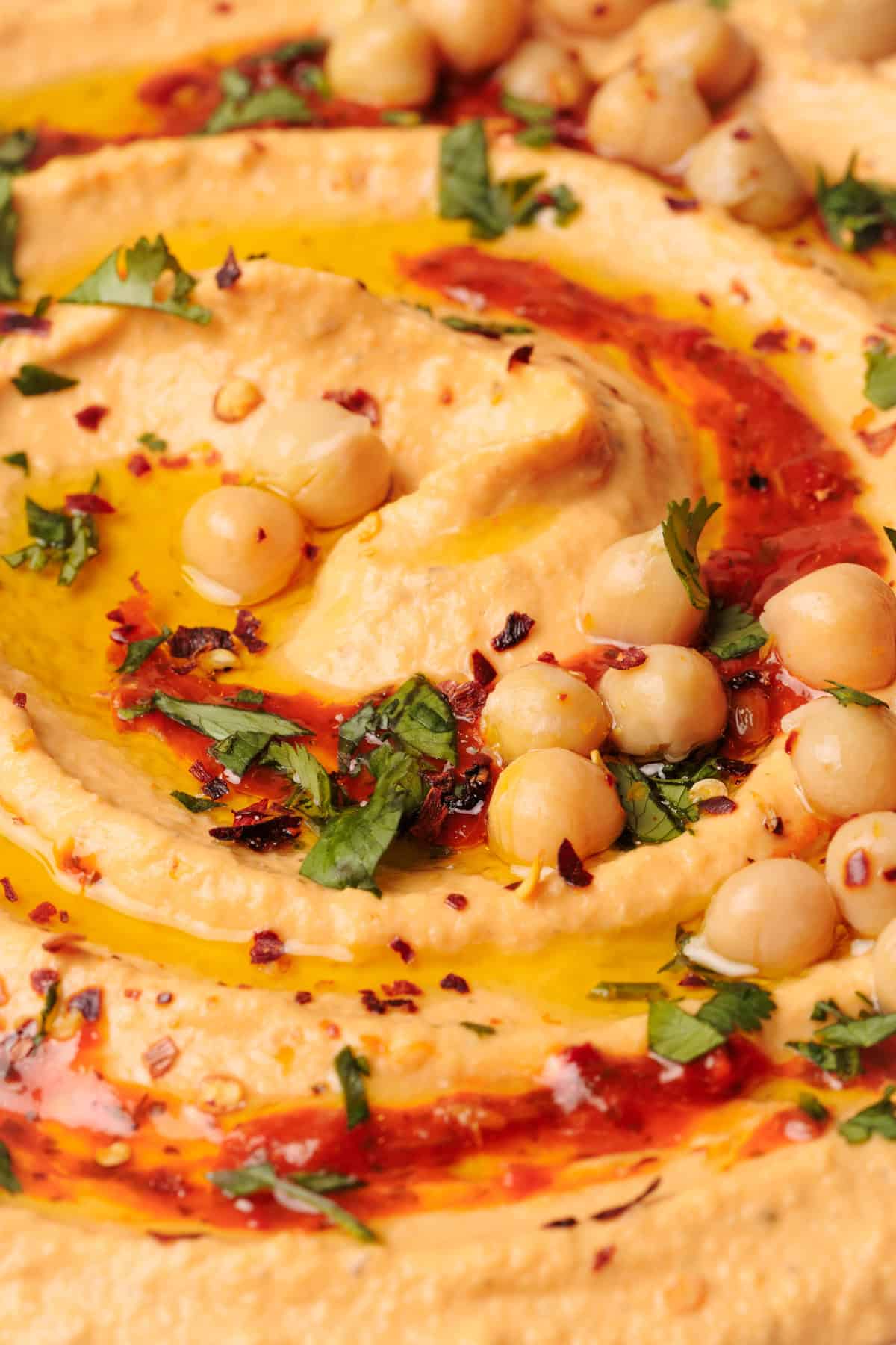 Close-up of creamy hummus with harissa topped with whole chickpeas, chopped parsley, olive oil, red pepper flakes, and paprika, creating a colorful, textured swirl.