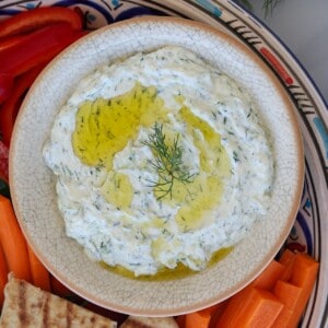 A bowl of tzatziki dip garnished with olive oil and dill, surrounded by sliced red peppers, carrots, and pita bread on a decorative plate.