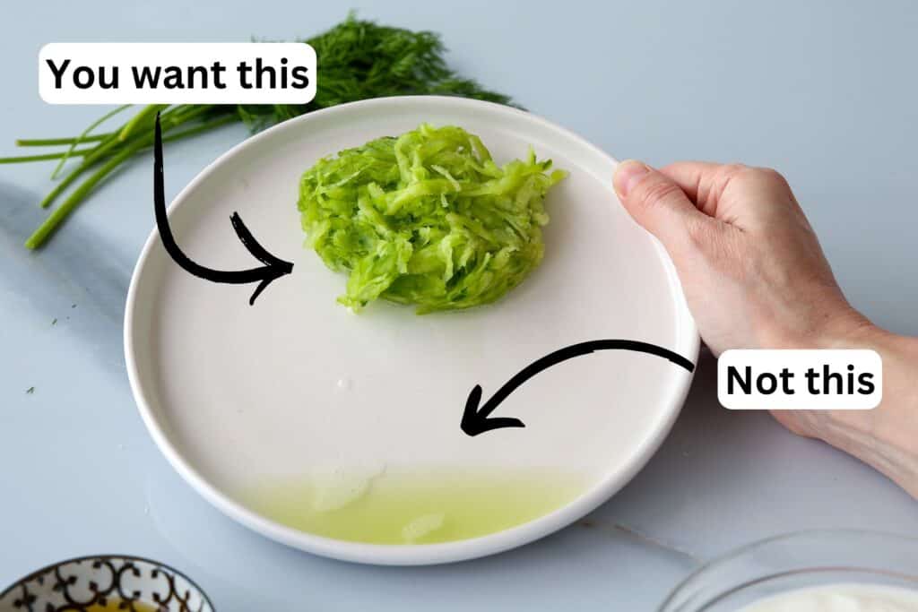 A hand holds a plate with grated cucumber and a small puddle of liquid. Arrows label the grated cucumber as “You want this” and the liquid as “Not this.” Fresh herbs are in the background.