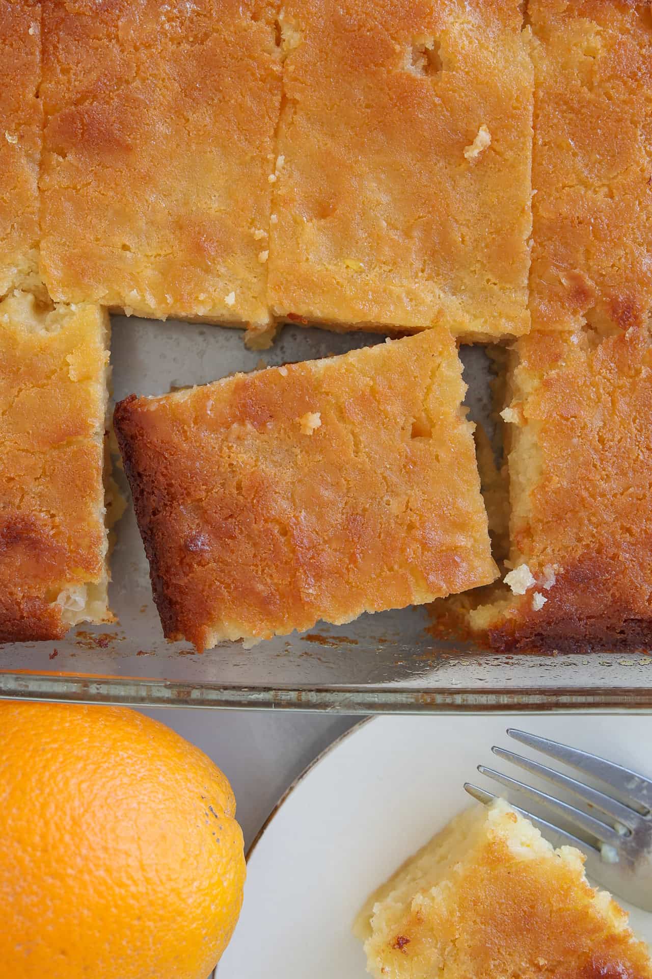 A tray of golden, sliced portokalopita (greek orange cake) with one piece pulled out, a fork on a plate with another cake slice, and an orange nearby.