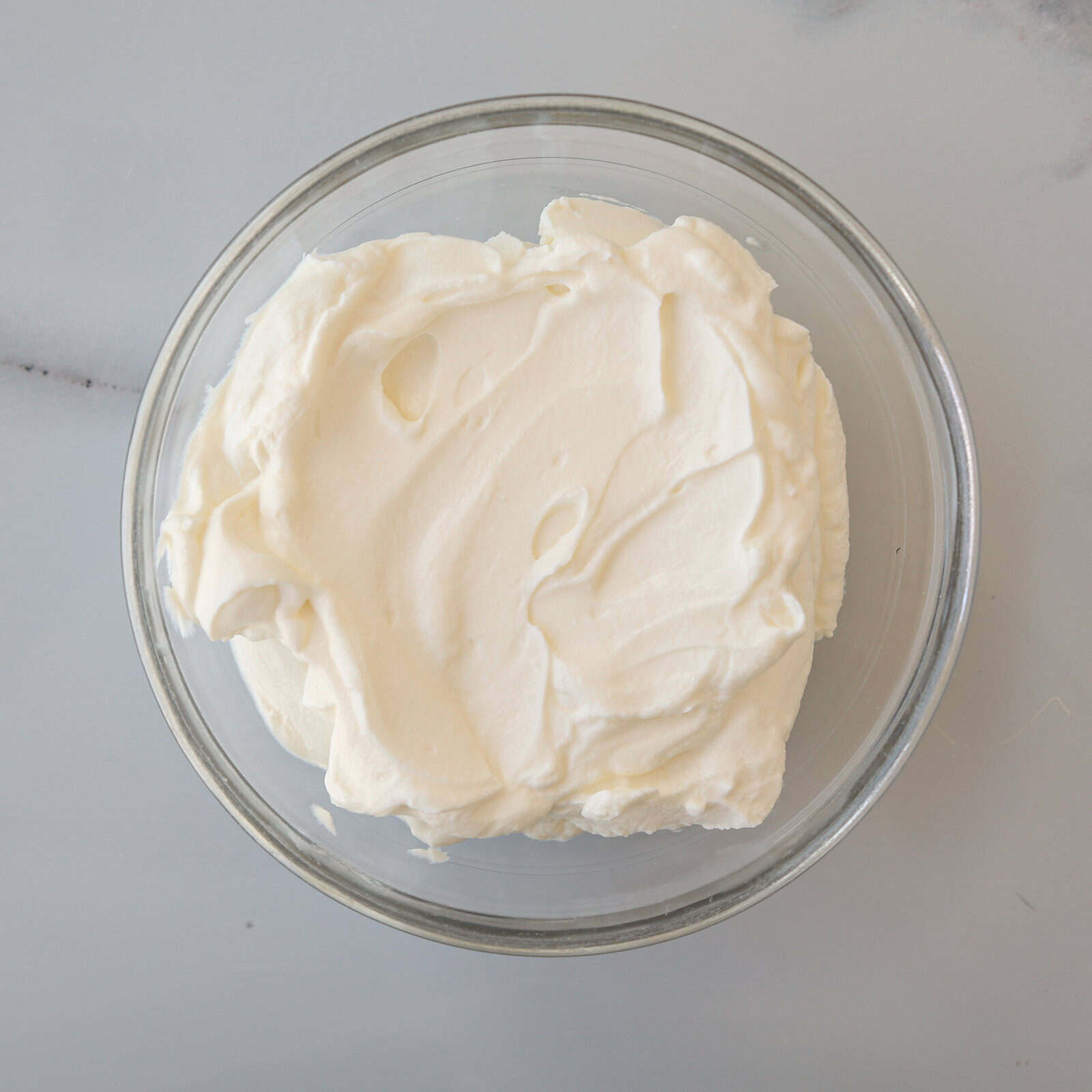 A clear glass bowl filled with creamy, thick Greek yogurt with a 10% fat content sits on a light gray marble surface.