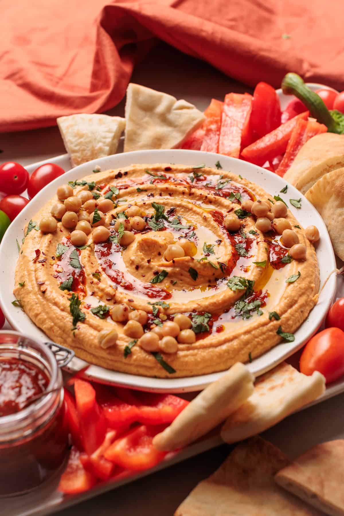 A plate of harissa hummus spread topped with chickpeas, herbs, olive oil, and spices, surrounded by pita bread slices, red bell pepper strips, and cherry tomatoes on a table with an orange cloth in the background.
