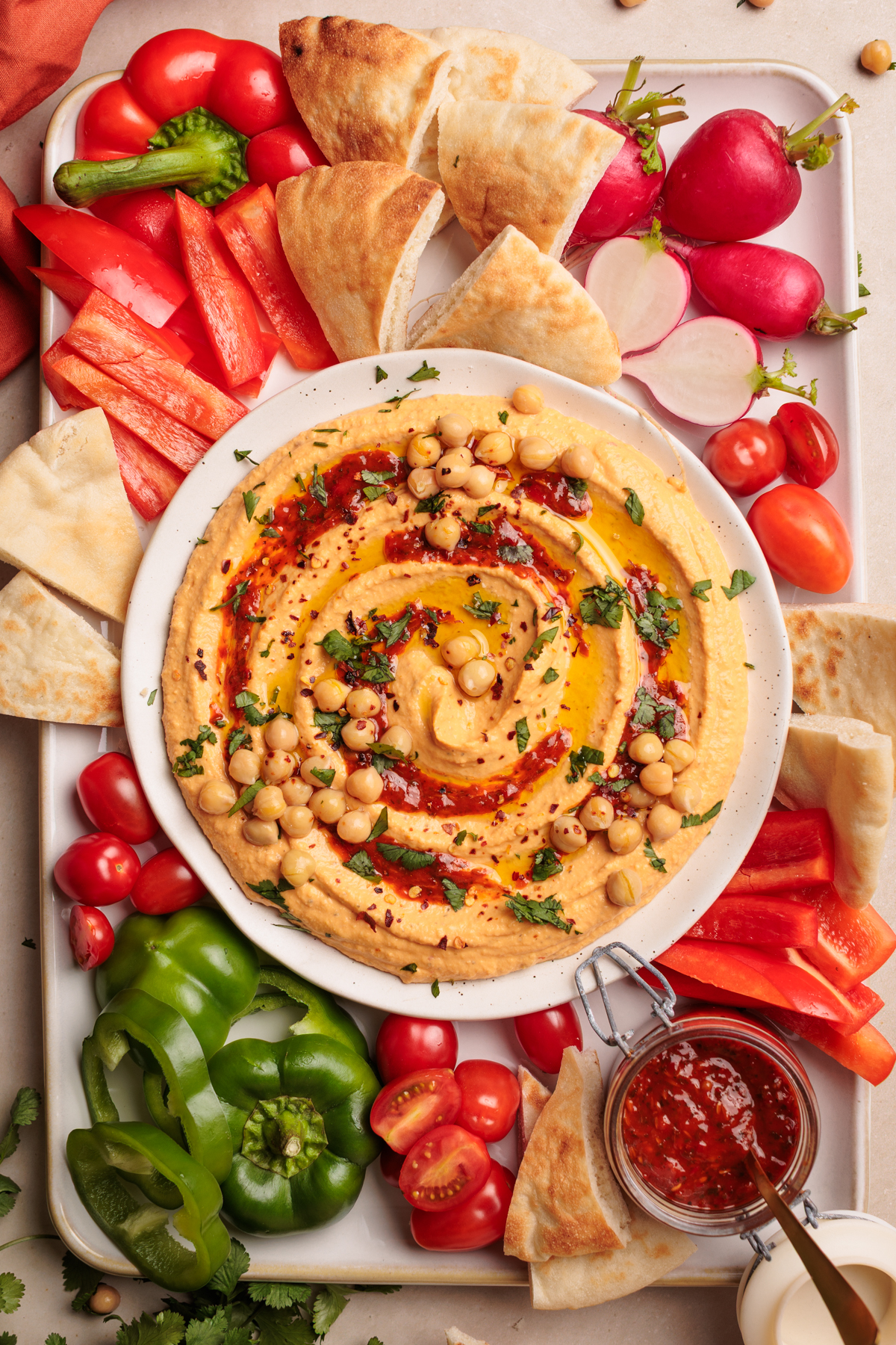 A plate of harissa hummus garnished with chickpeas, herbs, oil, and spices is surrounded by pita bread, bell peppers, cherry tomatoes, and radishes. A jar of homemade harissa paste is also present on the platter.