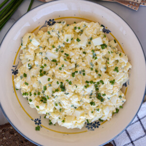 A bowl of creamy egg salad with cottage cheese garnished with chopped chives, served on a white plate with a floral pattern.