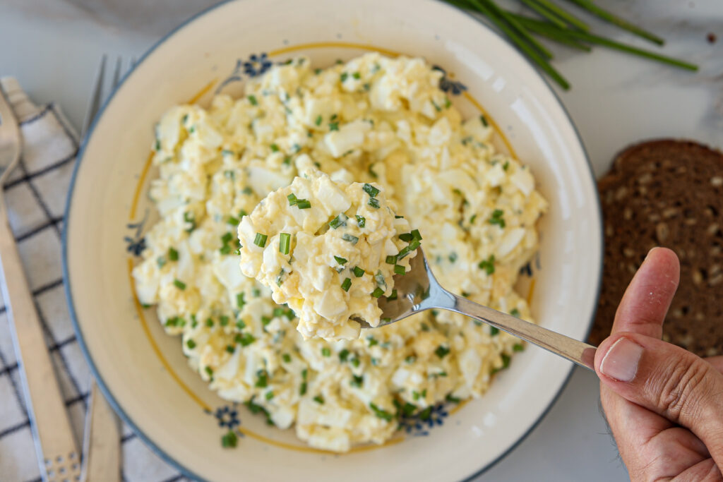 A hand holds a forkful of creamy egg salad garnished with chopped chives above a plate filled with more egg salad. Slices of bread and fresh chives are nearby.