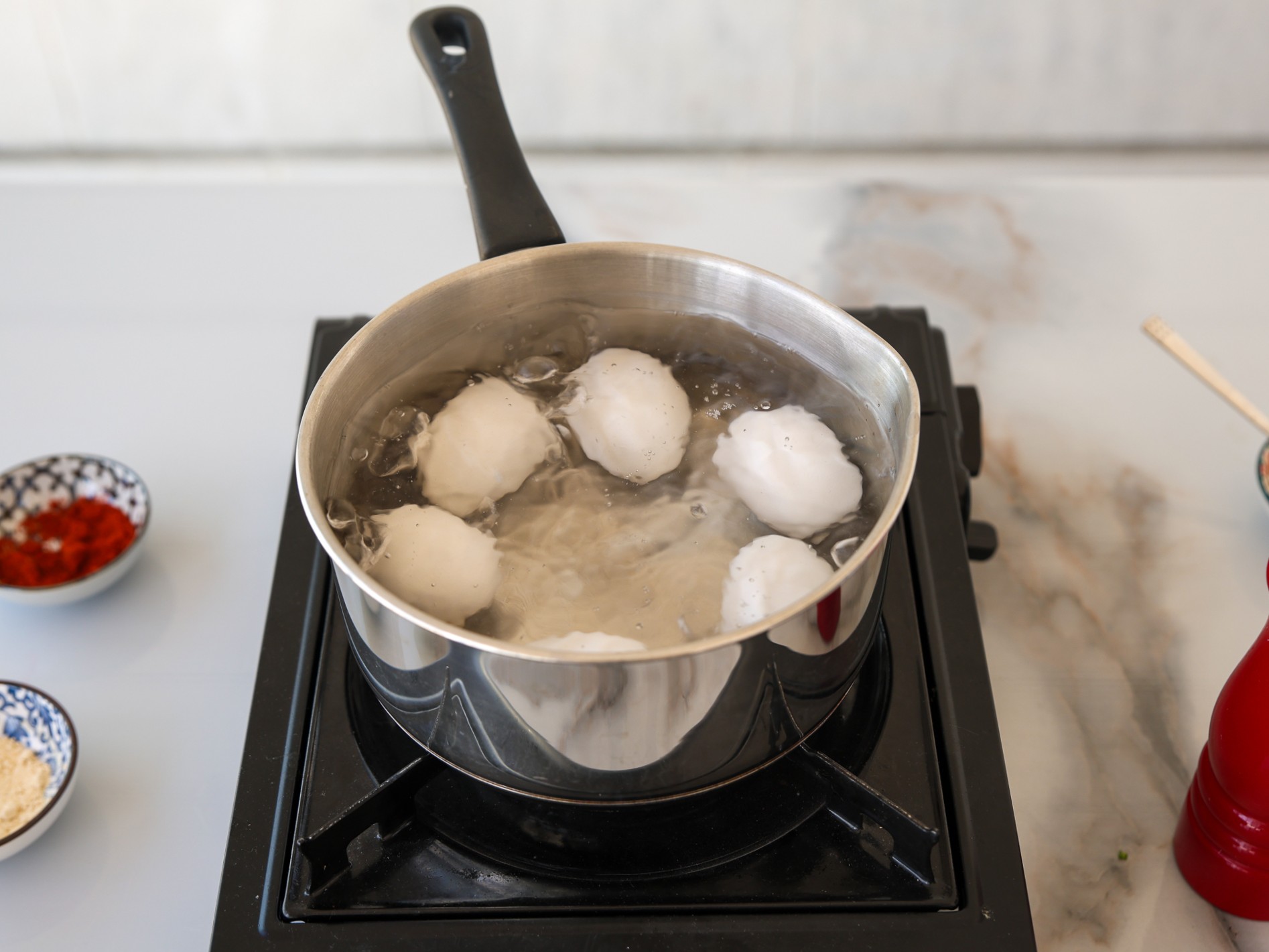 A stainless steel pot with six eggs boiling in water sits on a black stovetop. 