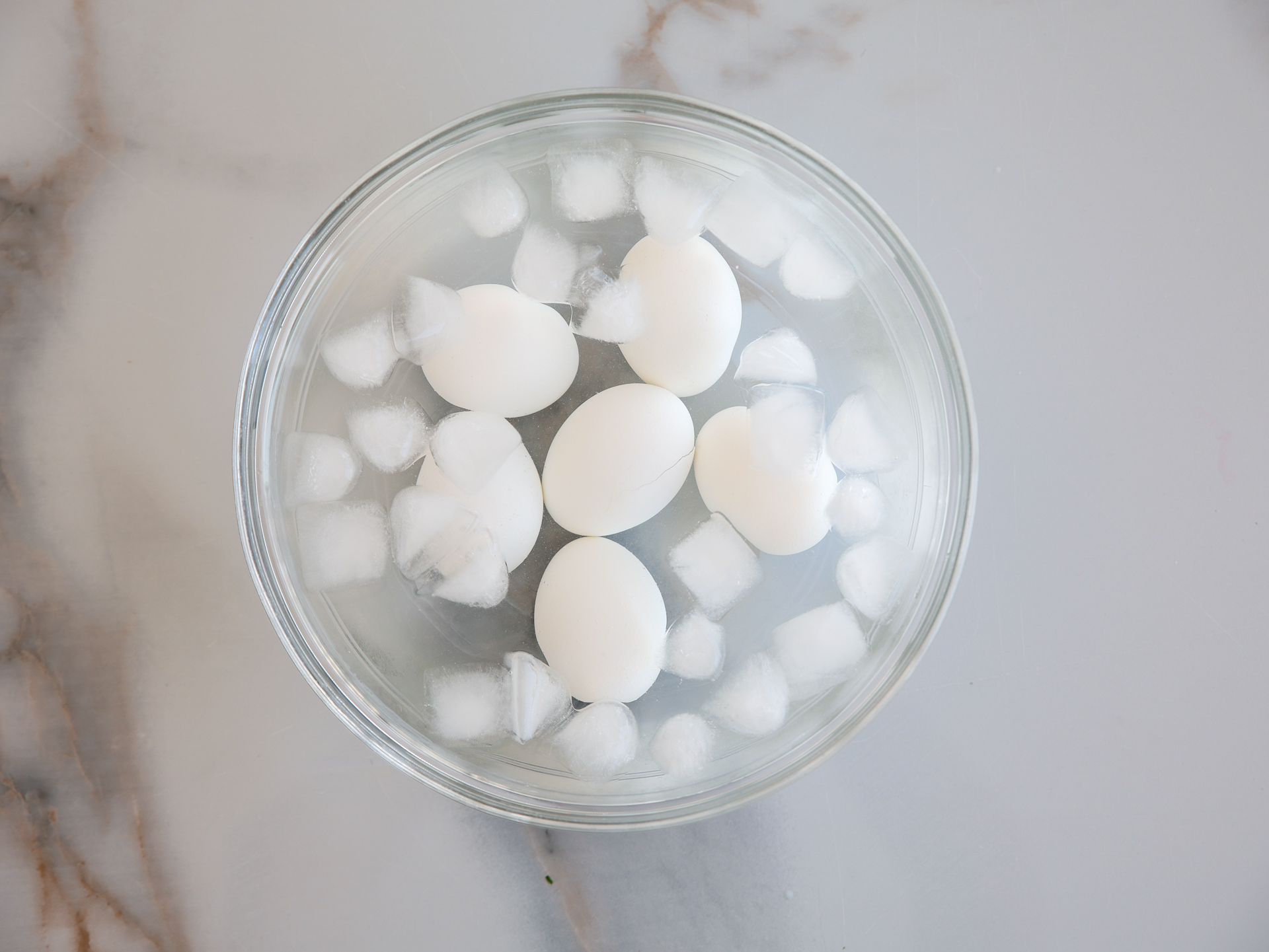 A glass bowl filled with ice water and six hard-boiled eggs is placed on a light marble surface.
