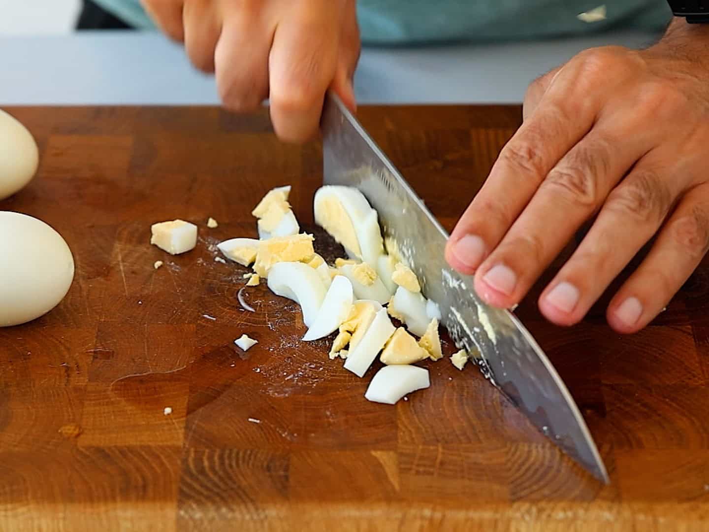 A person chopping a hard-boiled egg with a large knife on a wooden cutting board. Two uncut eggs are on the board.