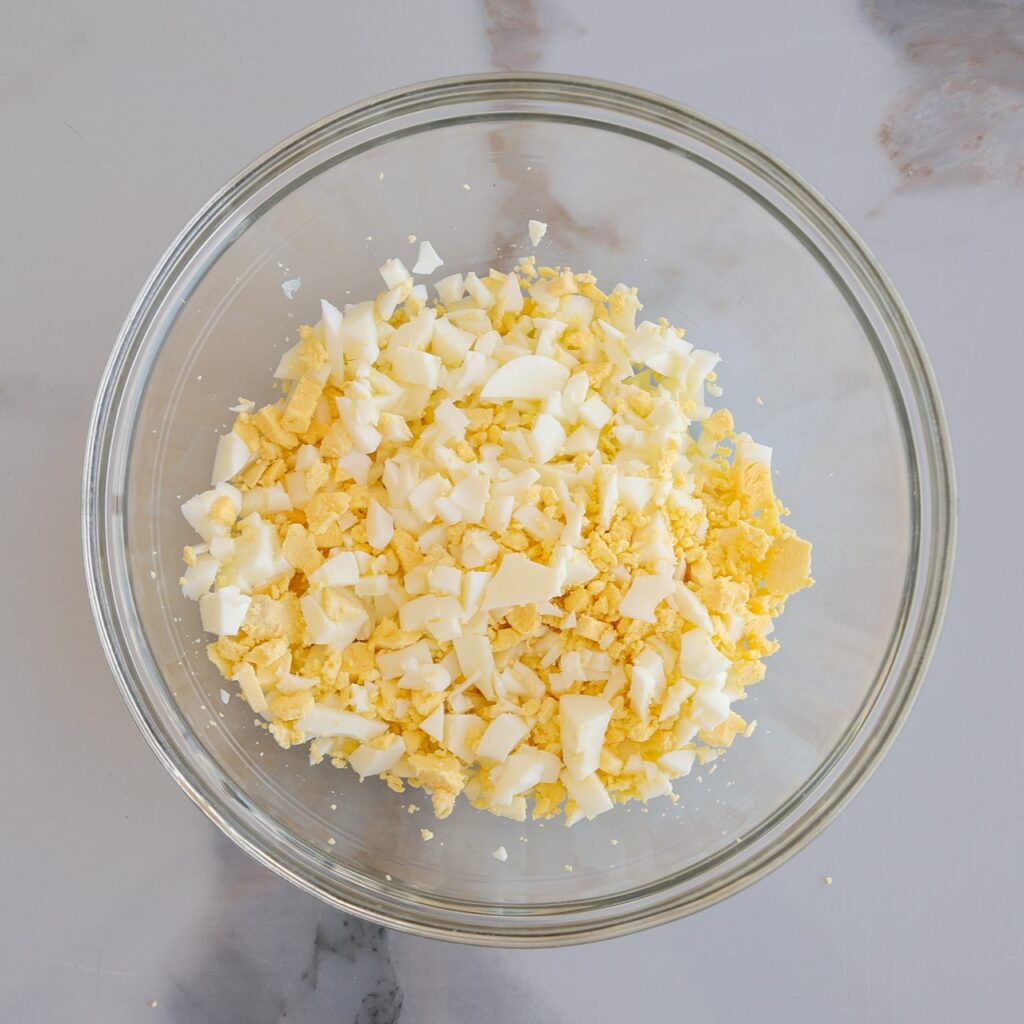 A clear glass bowl filled with finely chopped hard-boiled eggs, showing yellow yolks and white egg whites, sits on a light gray marble surface.