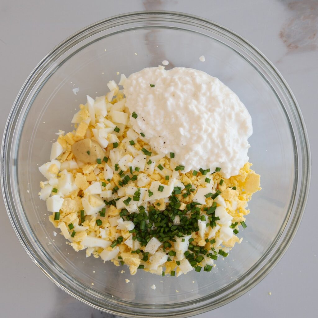 A glass bowl filled with chopped hard-boiled eggs, cottage cheese, fresh chopped chives, and seasoning on a light-colored surface.