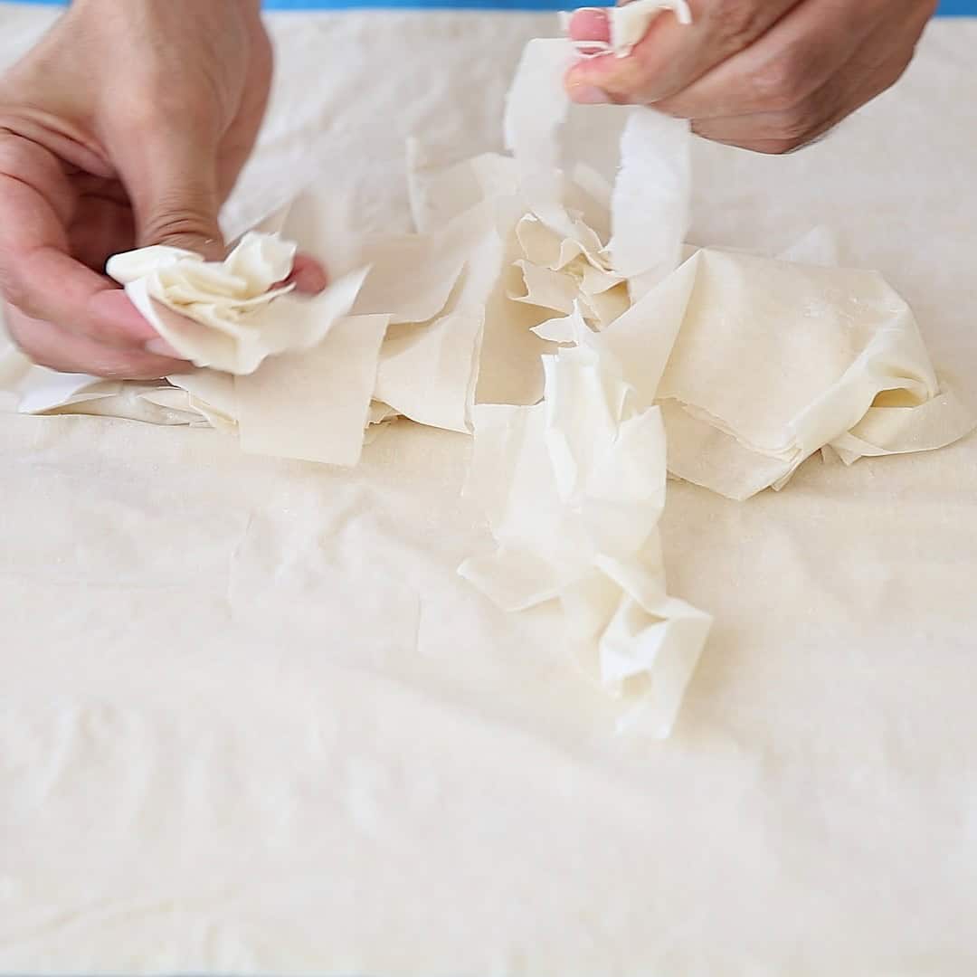 Two hands tearing sheets of phyllo dough over a flat surface covered with more phyllo sheets.