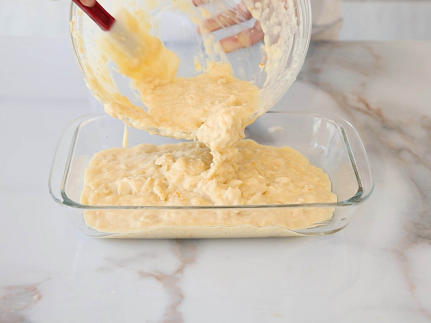A person pours thick, portokalopita batter from a glass bowl into a rectangular glass baking dish on a marble counter.
