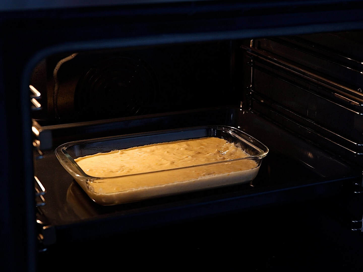 A glass baking dish filled with Greek orange cake batter sits on a rack inside a lit oven, baking.