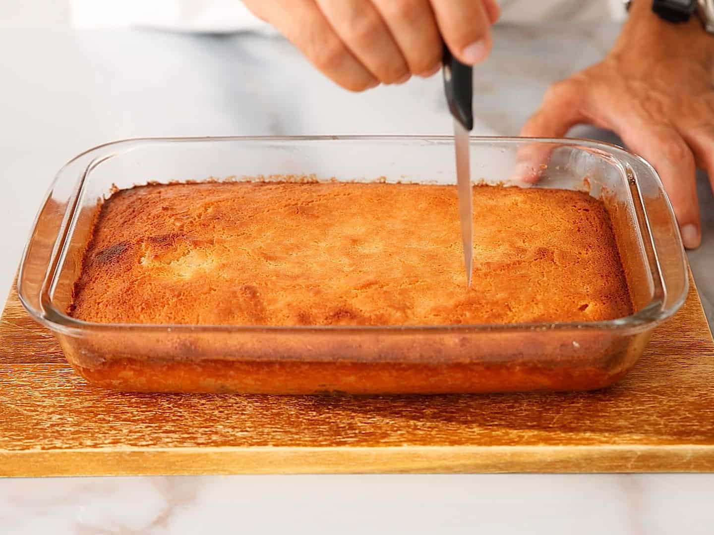 A person uses a knife to cut into a rectangular, golden-brown orange cake in a glass baking dish on a wooden board.