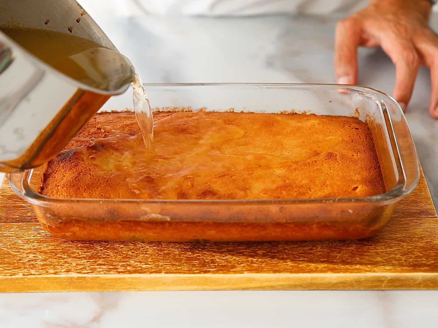 A hand pours clear syrup from a saucepan over a golden brown classic Greek orange cake in a glass baking dish on a wooden board.