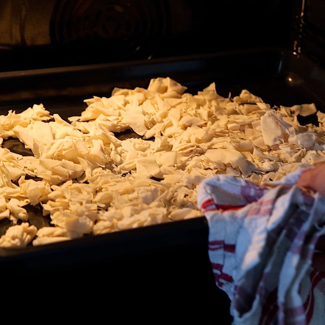 Chopped phyllo pieces spread on a baking tray inside an oven, with a person’s hand holding a red and white towel in the foreground.