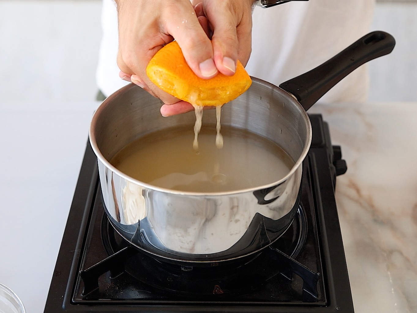 A person squeezes juice from an orange half into a pot of liquid on a stovetop, with juice visibly dripping into the pot.