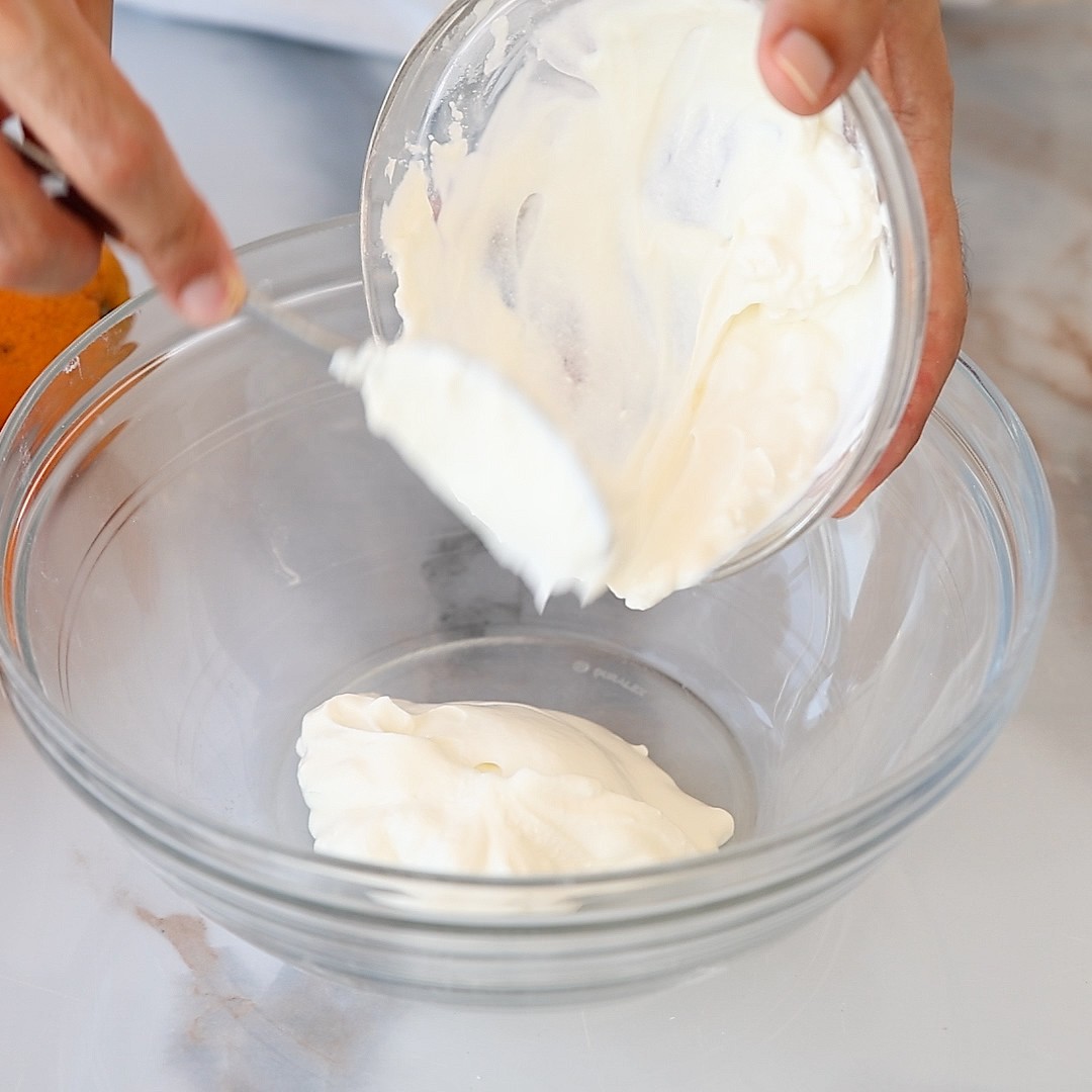 A person is spooning white creamy Greek yogurt from a small glass bowl into a larger clear mixing bowl on a marble countertop.